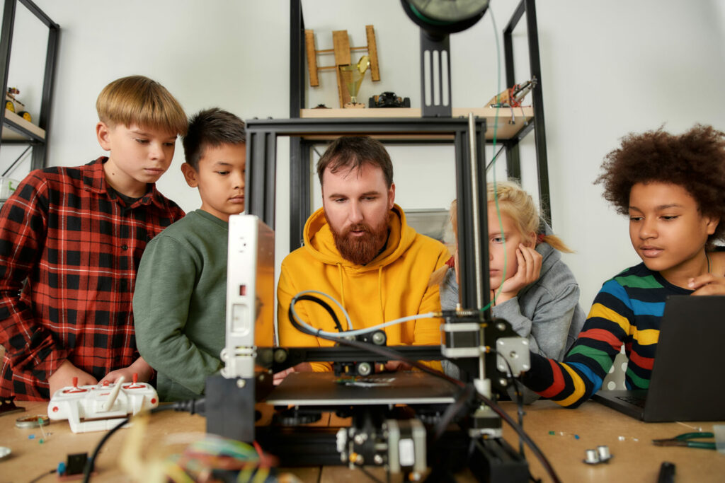 Experience digital innovation. Group of smart kids listening to their male teacher while looking at 3d printer and plastic detail prototype at robotics school lesson
